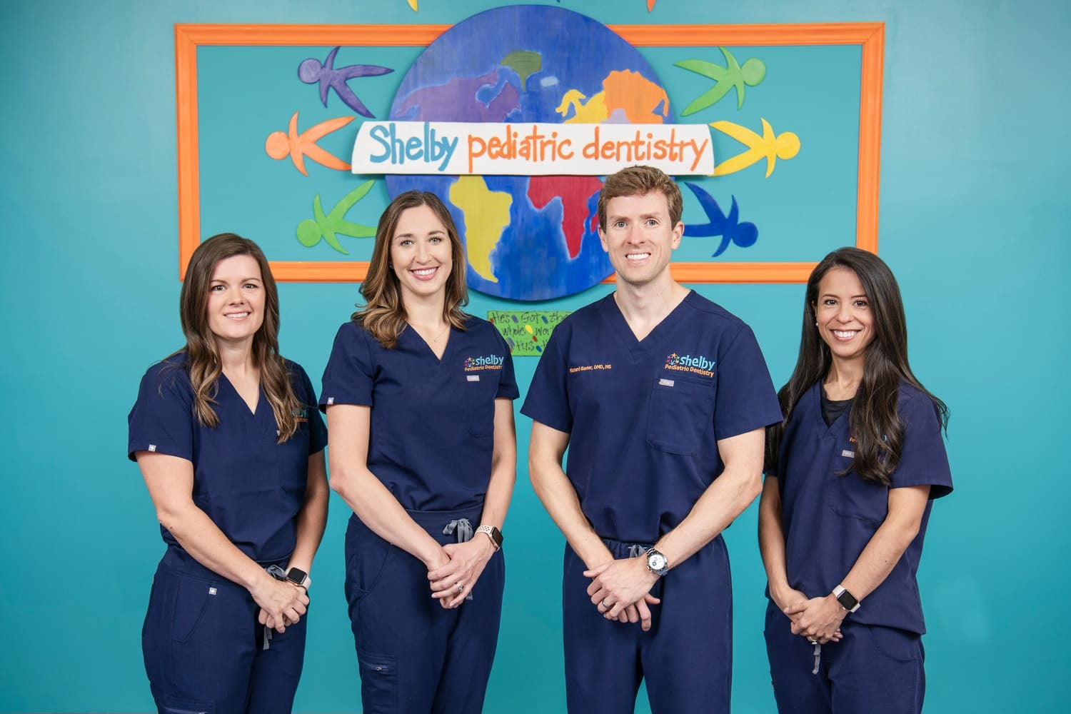 Four team members in navy scrubs pose in front of Shelby Pediatric Dentistry mural in Pelham, AL
