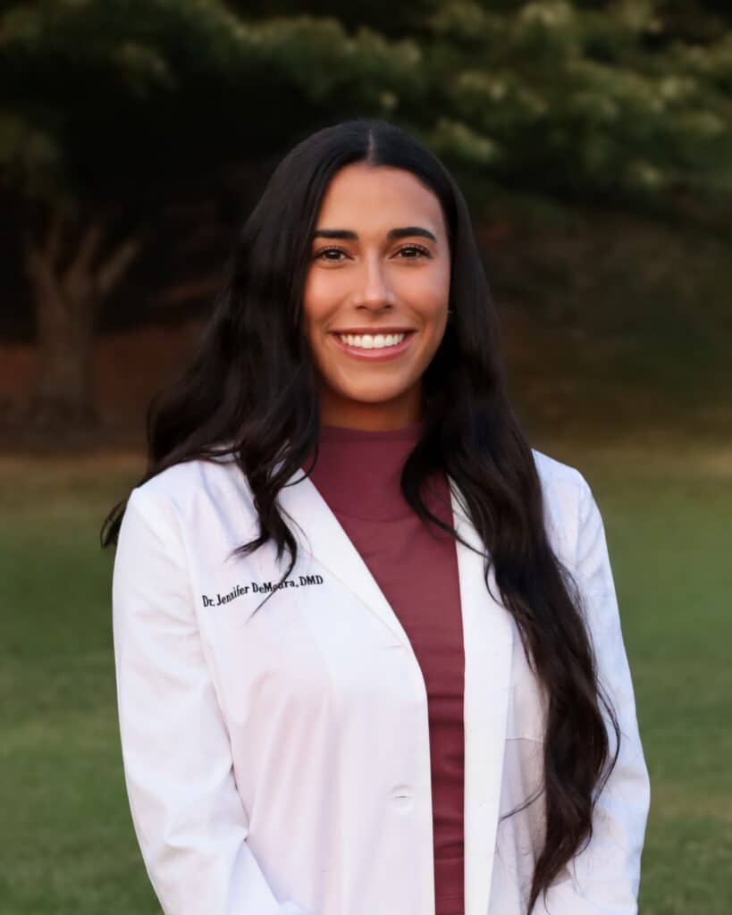 Dr. Jennifer DeMoura, DMD, smiling outdoors in a white dental coat