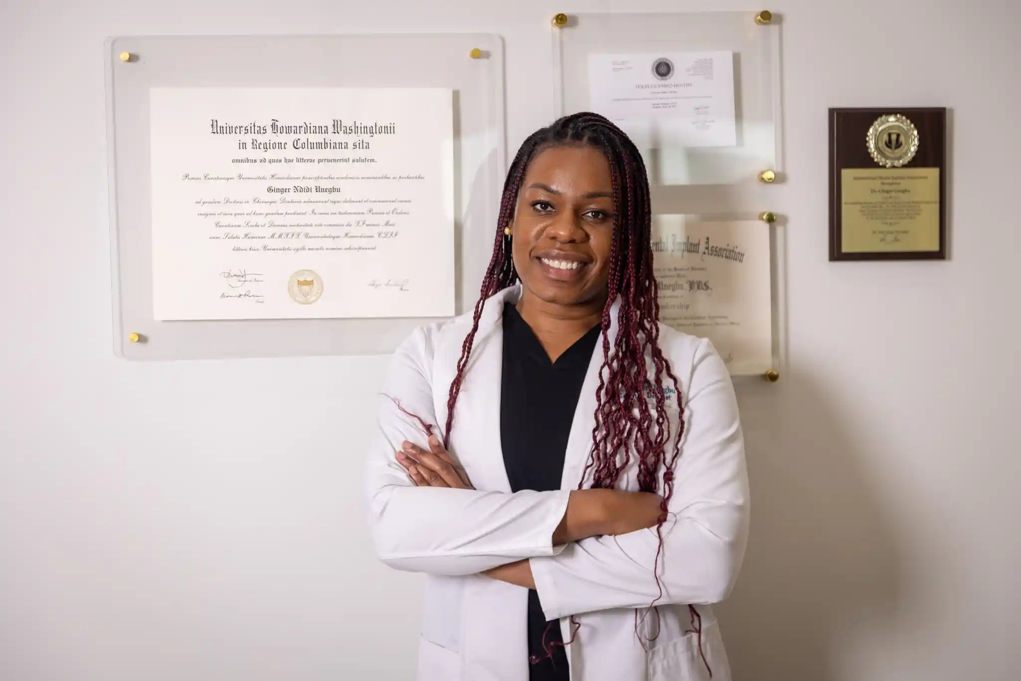 Dentist Dr. Ginger Unegbu stands smiling in front of her diplomas and certifications on the wall