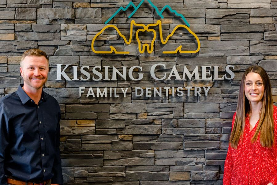 Two team members smiling in front of the Kissing Camels Family Dentistry stone wall sign in Colorado Springs