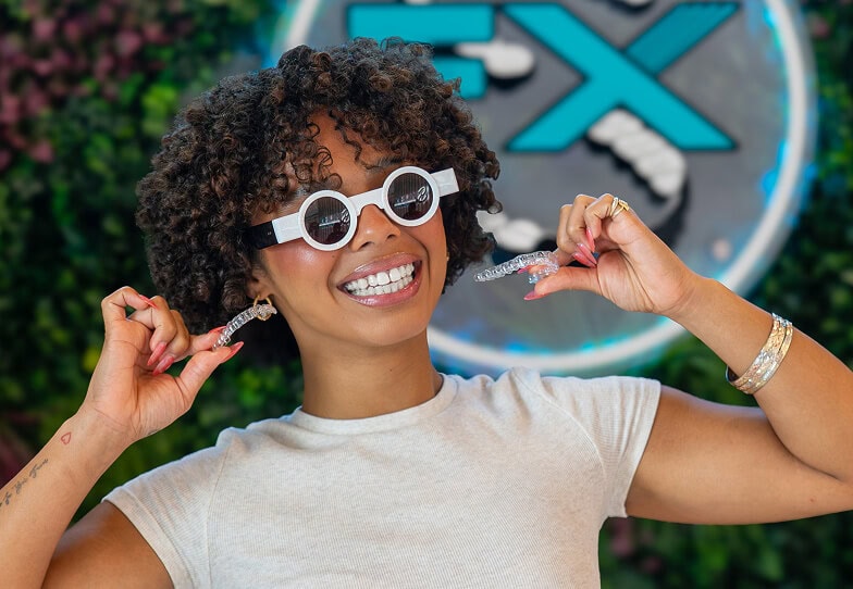 Smiling patient wearing novelty sunglasses holding clear aligners in front of SMILE-FX logo