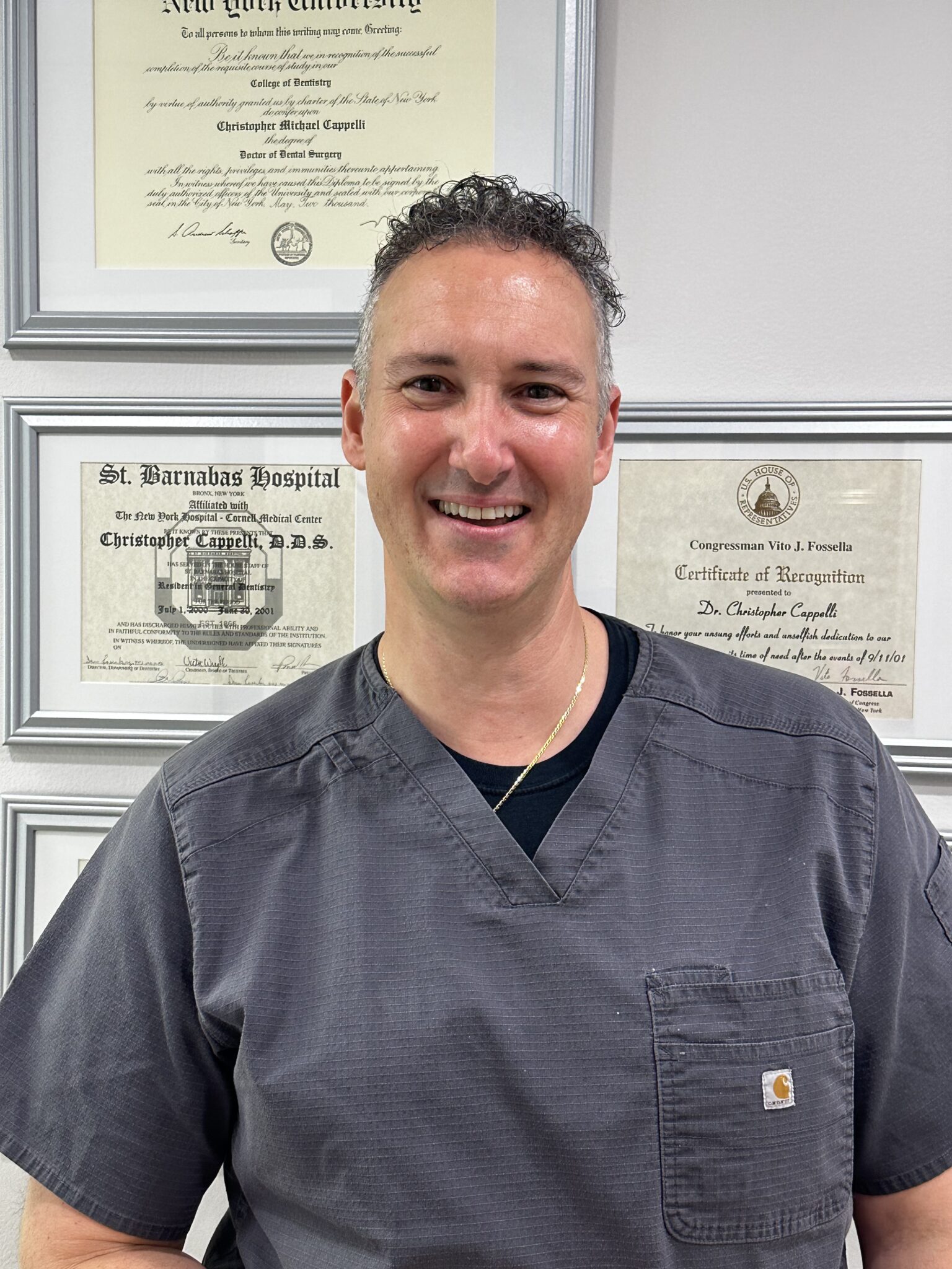 Dr. Christopher Cappelli smiling in front of his dental degree and hospital certificates at the practice