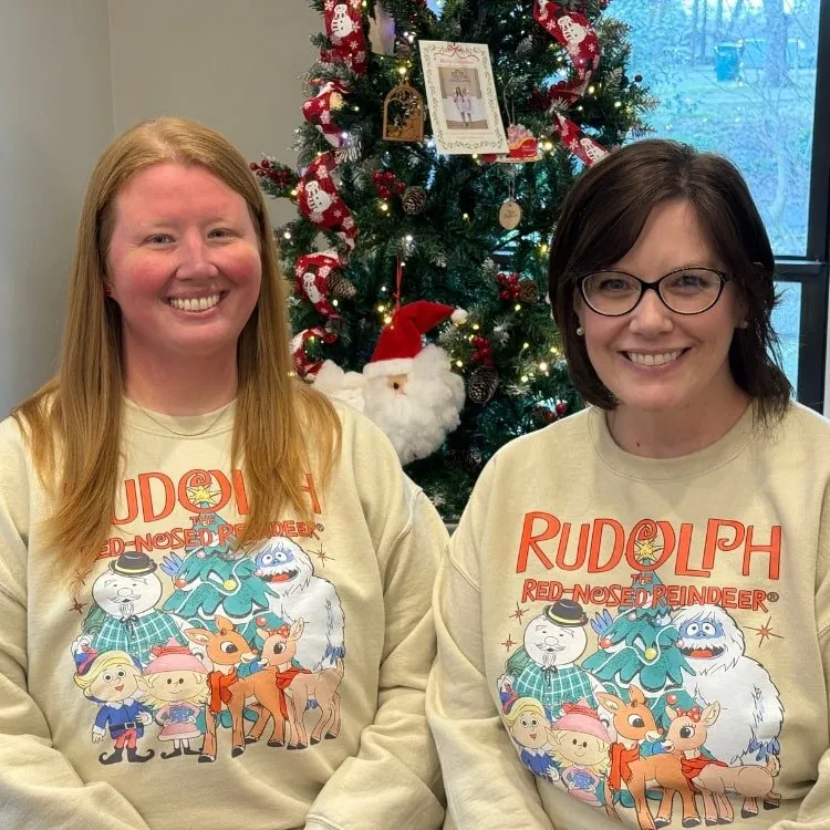 Two team members at Stone Dental Clinic in Wiggins, MS wearing matching holiday sweatshirts by a Christmas tree