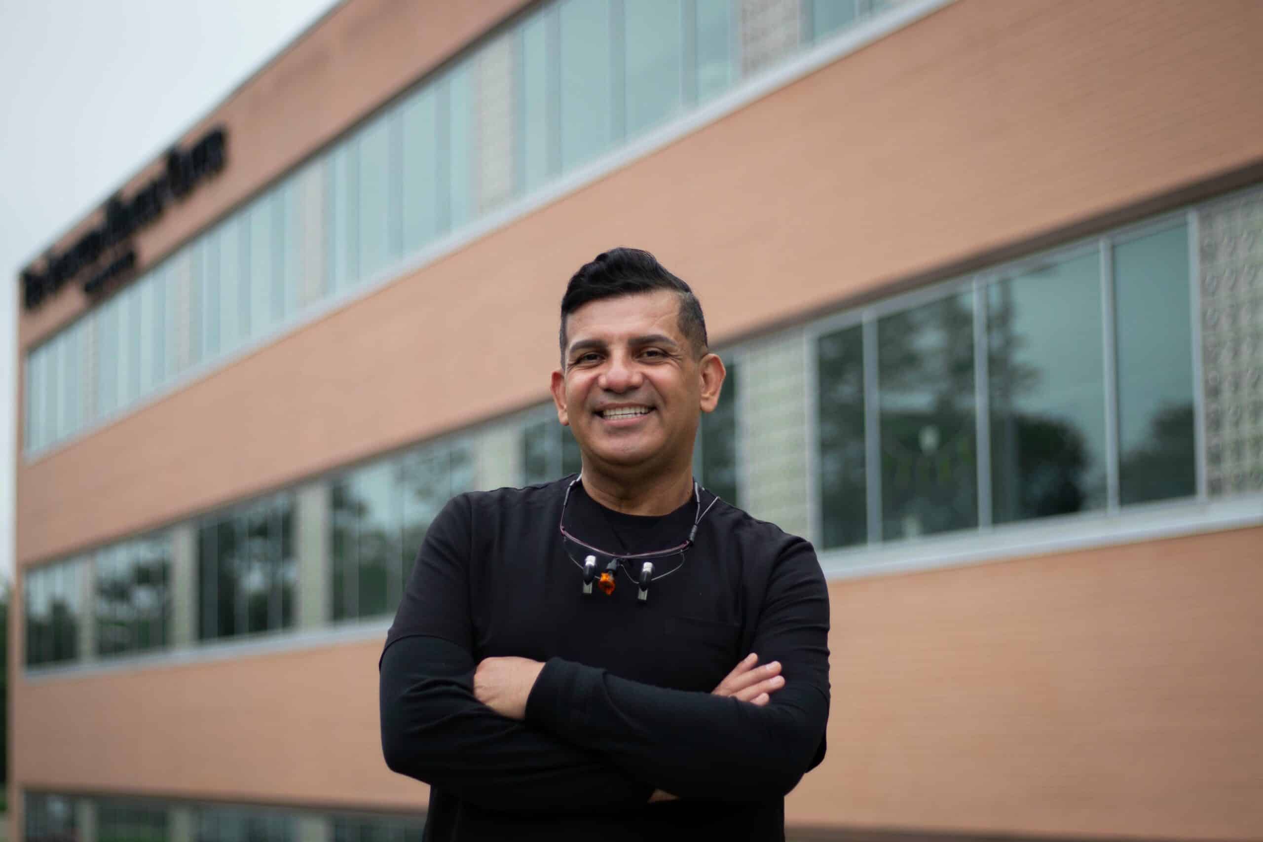 Team member in black shirt with arms crossed smiling outside the dental office building