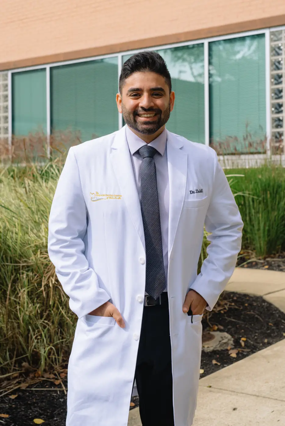 Dr. Zaidi in a white coat smiling outside The Advanced Dental Center building